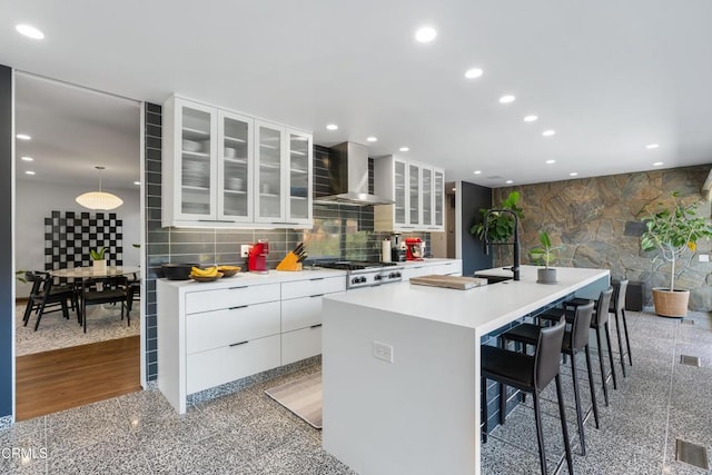 kitchen with hanging light fixtures, a kitchen island with sink, a breakfast bar area, white cabinets, and wall chimney exhaust hood