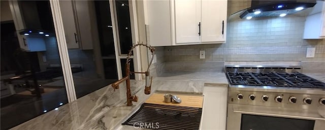 kitchen featuring wall chimney range hood, white cabinetry, stainless steel gas stove, light stone countertops, and decorative backsplash