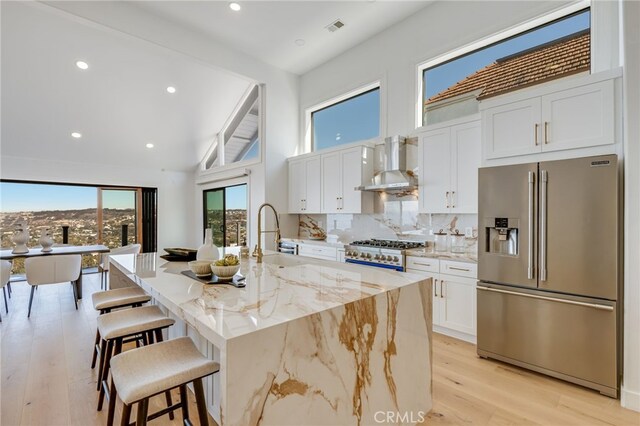 kitchen with white cabinetry, high end fridge, a center island with sink, and wall chimney exhaust hood