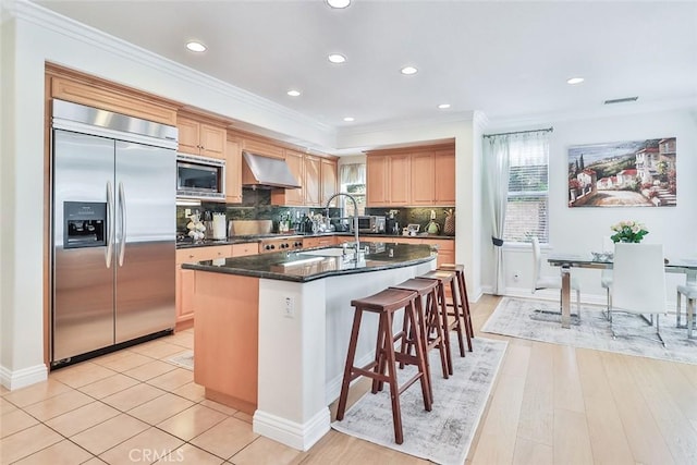 kitchen with an island with sink, dark stone counters, wall chimney range hood, crown molding, and built in appliances