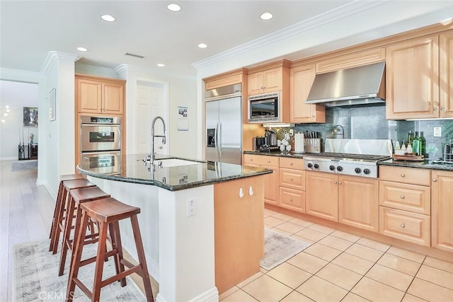 kitchen with wall chimney range hood, dark stone countertops, sink, built in appliances, and a kitchen island with sink