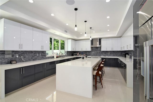 kitchen with pendant lighting, a raised ceiling, an island with sink, white cabinets, and wall chimney exhaust hood