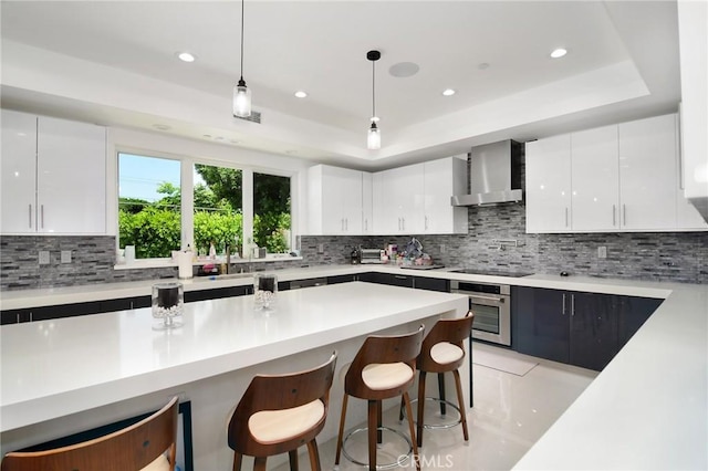 kitchen with decorative light fixtures, wall chimney range hood, oven, a tray ceiling, and white cabinetry