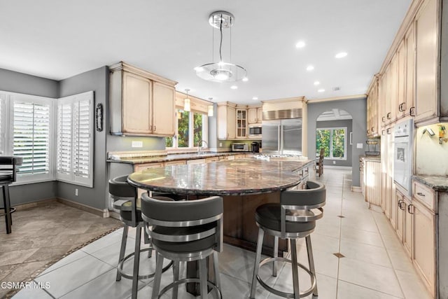 kitchen with dark stone countertops, a center island, pendant lighting, built in appliances, and light brown cabinets