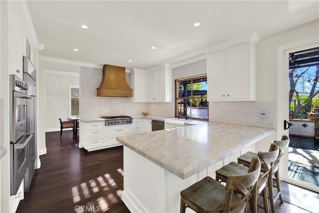 kitchen featuring custom range hood, appliances with stainless steel finishes, white cabinets, a sink, and a peninsula