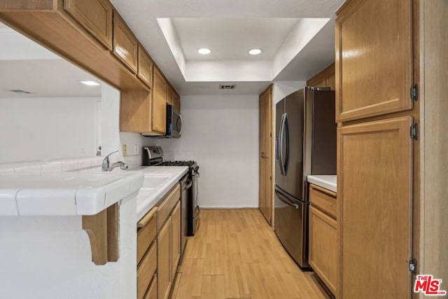 kitchen with tile countertops, sink, a tray ceiling, light wood-type flooring, and stainless steel appliances