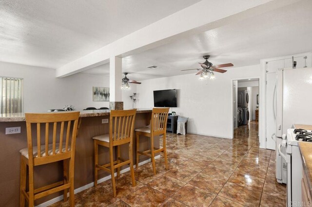 kitchen featuring stacked washer and clothes dryer, a kitchen bar, ceiling fan, white gas stove, and white cabinets