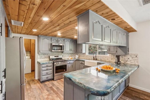 kitchen with washer / dryer, kitchen peninsula, appliances with stainless steel finishes, gray cabinetry, and light hardwood / wood-style flooring