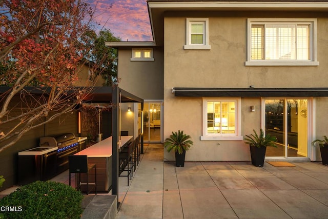 back of property at dusk with outdoor dry bar, a patio area, and stucco siding