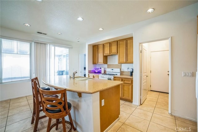 kitchen with sink, light tile patterned flooring, a kitchen breakfast bar, an island with sink, and white gas range