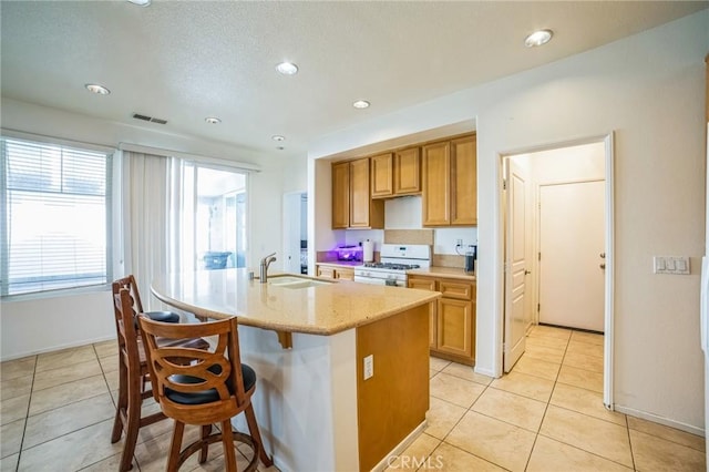 kitchen with white gas range, recessed lighting, light tile patterned flooring, a kitchen island with sink, and a sink