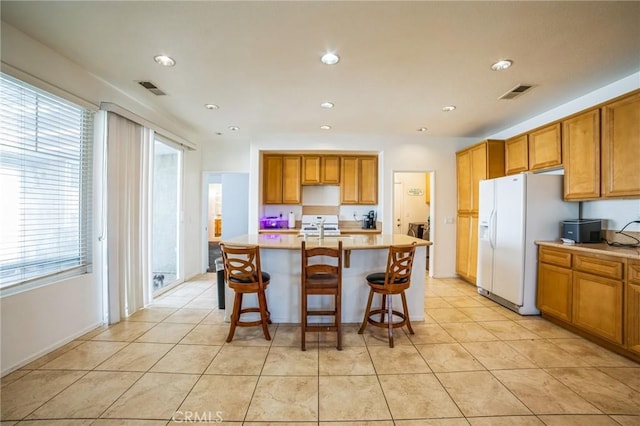 kitchen featuring light tile patterned floors, a center island with sink, white fridge with ice dispenser, a wealth of natural light, and a breakfast bar