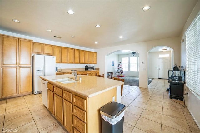 kitchen featuring arched walkways, light tile patterned floors, a kitchen island with sink, a sink, and white appliances