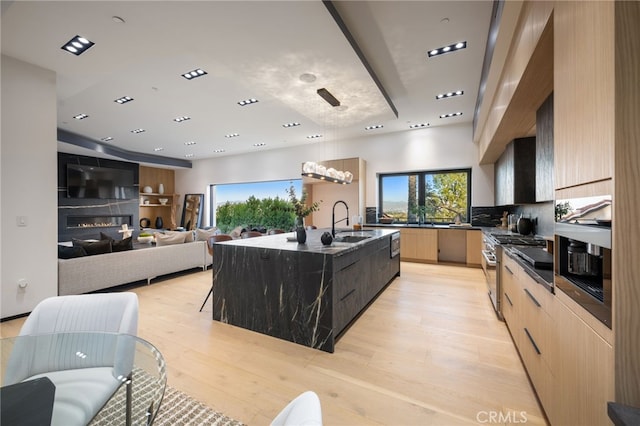 kitchen with sink, a kitchen island with sink, high end stainless steel range, light hardwood / wood-style floors, and light brown cabinetry