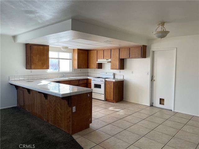 kitchen with sink, white gas range, kitchen peninsula, tile countertops, and light tile patterned floors