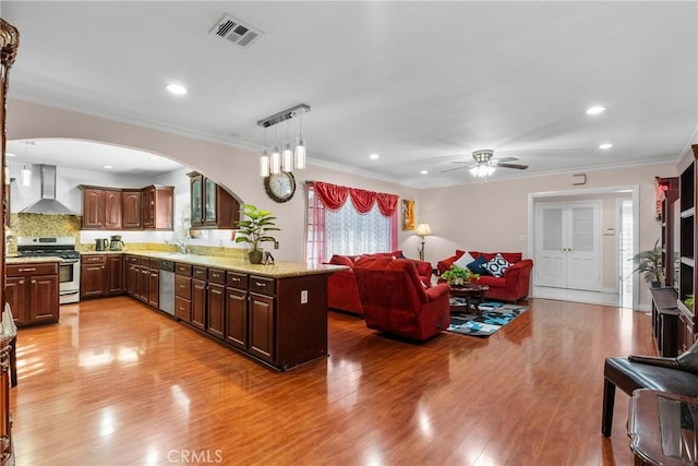 kitchen featuring hanging light fixtures, stainless steel appliances, wall chimney range hood, light wood-type flooring, and ornamental molding