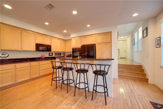 kitchen with light brown cabinets, a kitchen breakfast bar, light hardwood / wood-style floors, a center island with sink, and black appliances