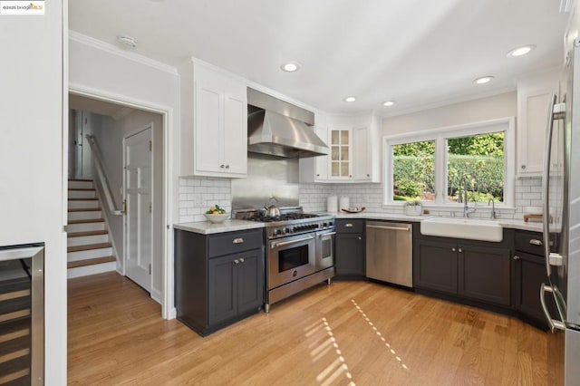 kitchen featuring wall chimney exhaust hood, stainless steel appliances, beverage cooler, white cabinetry, and sink