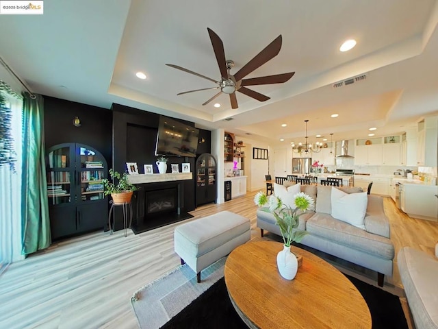 living room with ceiling fan with notable chandelier, a raised ceiling, and light wood-type flooring