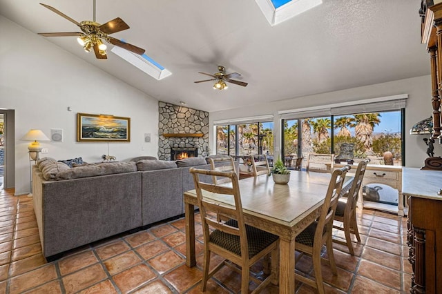 dining space with ceiling fan, vaulted ceiling with skylight, a stone fireplace, and tile patterned flooring