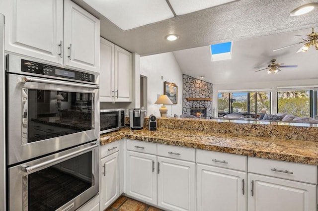 kitchen with white cabinets, appliances with stainless steel finishes, a fireplace, ceiling fan, and lofted ceiling with skylight