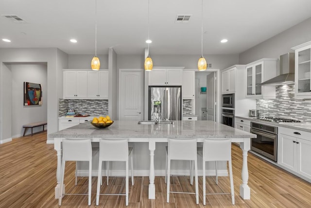 kitchen with decorative light fixtures, wall chimney range hood, stainless steel appliances, and a kitchen island with sink