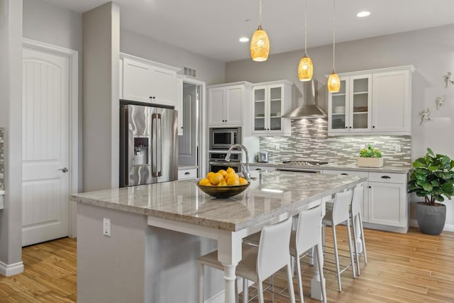 kitchen featuring white cabinets, wall chimney range hood, appliances with stainless steel finishes, and a center island with sink