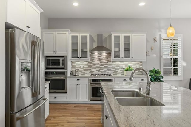 kitchen with light stone countertops, appliances with stainless steel finishes, white cabinets, wall chimney range hood, and sink