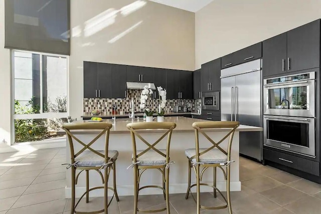 kitchen with light tile patterned floors, a kitchen bar, tasteful backsplash, a towering ceiling, and built in appliances