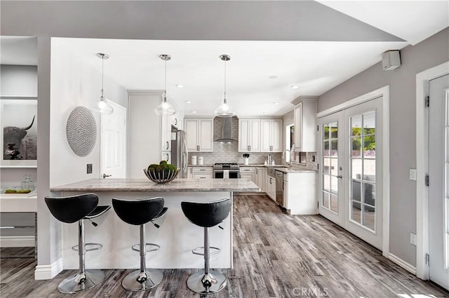 kitchen with hanging light fixtures, appliances with stainless steel finishes, white cabinets, french doors, and wall chimney exhaust hood