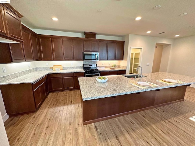 kitchen with an island with sink, stainless steel appliances, light hardwood / wood-style flooring, light stone counters, and sink