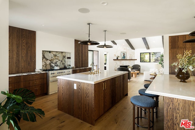 kitchen featuring high end stainless steel range oven, a stone fireplace, a kitchen island with sink, light wood-type flooring, and vaulted ceiling with beams