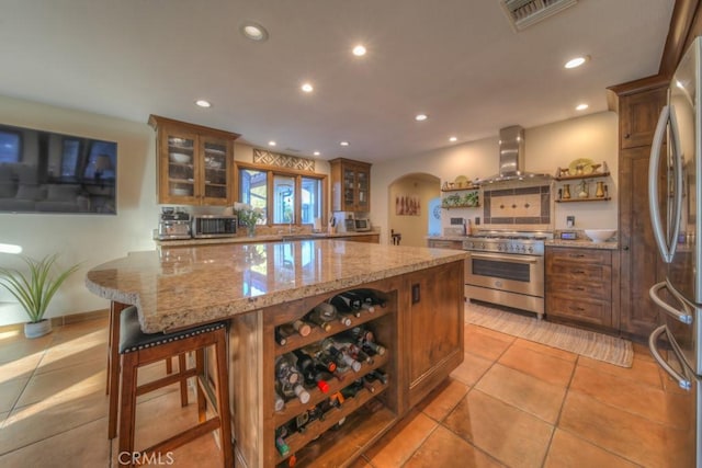 kitchen featuring a kitchen bar, stainless steel appliances, light tile patterned floors, wall chimney exhaust hood, and light stone counters