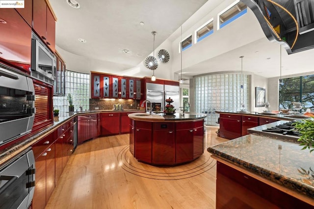 kitchen featuring decorative light fixtures, built in appliances, an island with sink, sink, and light hardwood / wood-style flooring