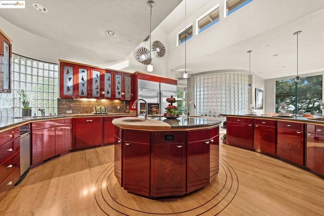 kitchen featuring stainless steel built in refrigerator, tasteful backsplash, light hardwood / wood-style floors, an island with sink, and hanging light fixtures