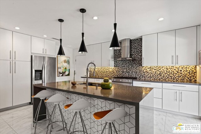 kitchen featuring decorative light fixtures, stainless steel appliances, an island with sink, wall chimney range hood, and a kitchen bar