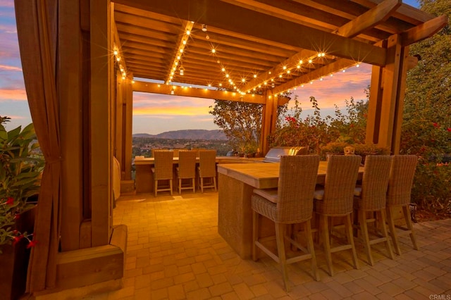 patio terrace at dusk featuring exterior kitchen, exterior bar, a mountain view, and a pergola