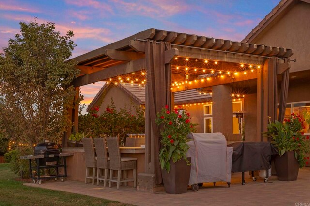 patio terrace at dusk featuring grilling area, a pergola, and exterior bar