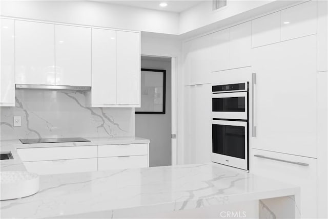 kitchen with light stone countertops, multiple ovens, black electric cooktop, exhaust hood, and white cabinetry