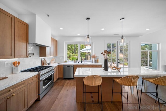 kitchen with tasteful backsplash, light hardwood / wood-style floors, hanging light fixtures, appliances with stainless steel finishes, and wall chimney exhaust hood