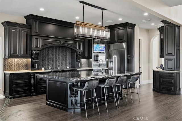 kitchen with decorative backsplash, sink, a kitchen island with sink, dark wood-type flooring, and stainless steel built in fridge