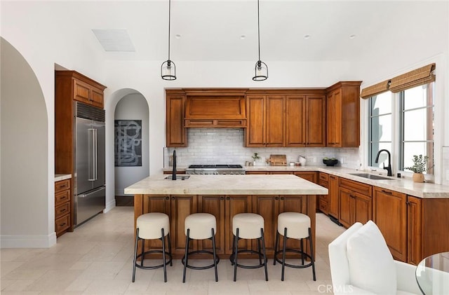 kitchen featuring backsplash, a kitchen island with sink, decorative light fixtures, built in fridge, and sink