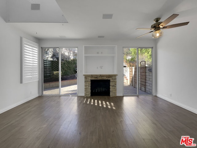 unfurnished living room with ceiling fan, built in shelves, dark wood-type flooring, and a stone fireplace