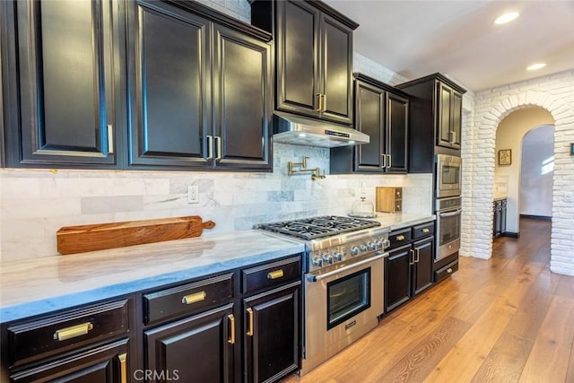 kitchen with brick wall, stainless steel appliances, tasteful backsplash, light wood-type flooring, and light stone counters