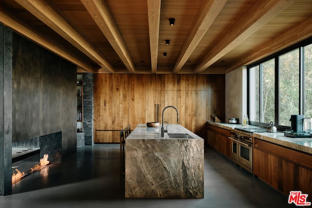 kitchen with wood walls, sink, a kitchen island with sink, light stone countertops, and range with two ovens