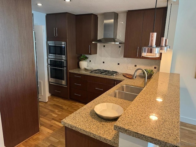 kitchen featuring kitchen peninsula, appliances with stainless steel finishes, wall chimney range hood, light stone countertops, and sink