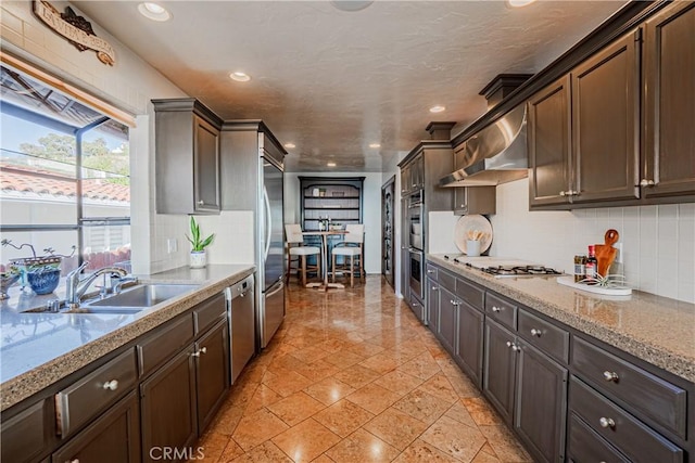 kitchen with appliances with stainless steel finishes, wall chimney range hood, dark brown cabinets, and sink