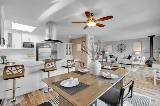 dining area featuring ceiling fan, sink, a wood stove, a skylight, and light hardwood / wood-style floors