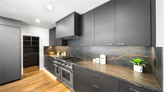 kitchen featuring double oven range, light hardwood / wood-style floors, decorative backsplash, dark stone counters, and wall chimney exhaust hood