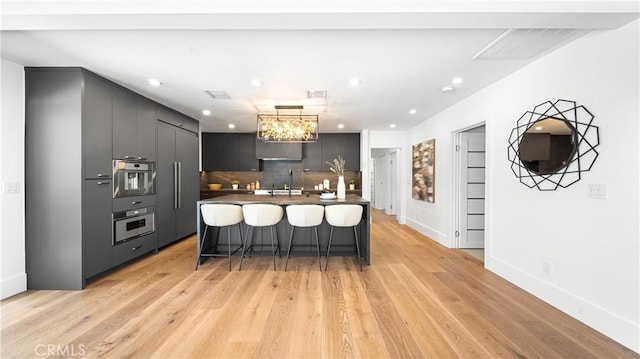 kitchen featuring a kitchen bar, an island with sink, wall oven, backsplash, and light hardwood / wood-style flooring